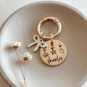 May include: A gold-colored keychain with a round charm engraved with the name "Charlie" and small seashell, starfish, and ornament designs. A white bow charm and a rose gold clasp complete the design. The keychain is on a neutral-colored tray.