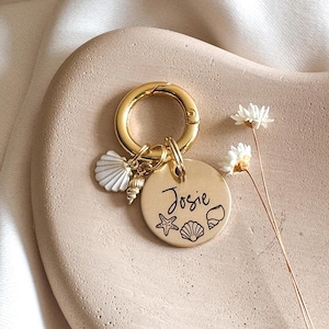 May include: A gold-colored keychain with a round tag and seashell charms. The tag is engraved with the name "Josie" and seashell illustrations. The keychain is on a light pink surface.