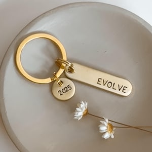 May include: Gold keychain with a small round charm engraved with the year 2025 and a larger rectangular charm engraved with the word "EVOLVE".