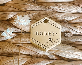 Könnte beinhalten: Goldenes sechseckiges Schild mit dem Wort "HONEY" in der Mitte eingraviert. Unter dem Wort befindet sich ein kleines Bienenmotiv. Das Schild hat oben ein Loch zum Anbringen.