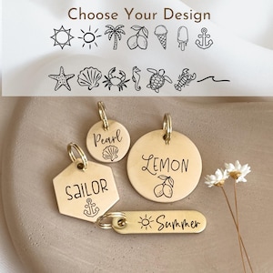 May include: A selection of brass pet tags and keychains with various designs. The tags include the words "Pearl", "Lemon", "Sailor", and "Summer", along with beach-themed illustrations like an anchor and sun.