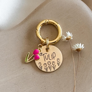 May include: A gold-colored pet tag with a round charm engraved with the word "Tulip" and tulip illustrations. A pink and green tulip charm hangs from the tag, attached to a gold clasp.