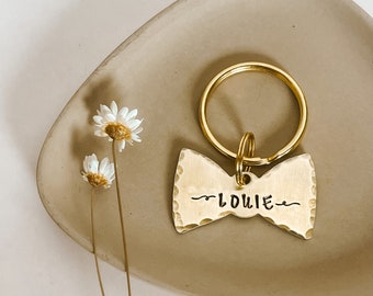 May include: Gold bowtie-shaped dog tag with the name "Louie" engraved on it. The tag is attached to a gold key ring.