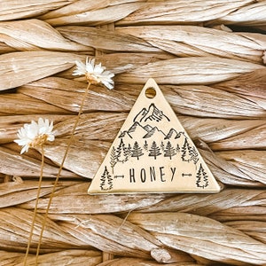 May include: A gold triangle-shaped tag with a mountain range and pine trees engraved on it. The text "HONEY" is engraved below the trees.