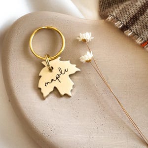May include: A gold-toned metal keychain featuring a maple leaf-shaped charm. The charm is engraved with the word maple in a black cursive script. A circular gold-toned split ring is attached to the top of the leaf charm by a small jump ring.