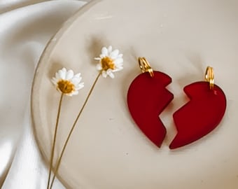 Puede incluir: Dos encantos de corazón de vidrio rojo con detalles dorados, rotos por la mitad, en un plato blanco con flores blancas.