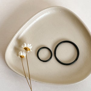 May include: Two black metal key rings, one larger than the other, are displayed on a beige ceramic tray. Dried white daisies are arranged on the tray.