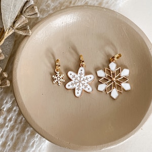 May include: Three gold snowflake charms with white enamel. The charms are on a light brown surface.