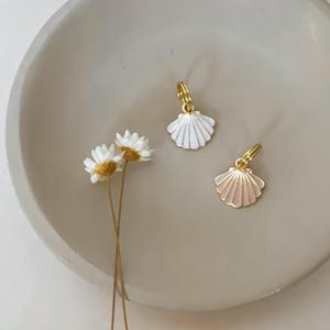 May include: Two seashell-shaped earrings with gold-colored hoops. One earring is white, and the other is pink. The earrings are displayed on a light-colored, round dish with a few small white flowers.