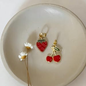 May include: Two gold-colored fruit charms: a red strawberry with green leaves and a pair of red cherries with green leaves. The charms are set on a beige, shallow dish, with a few small white flowers.