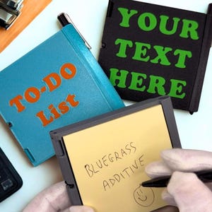 May include: A collection of small, square notebooks with pens. One notebook is blue with "TO-DO List" in orange. Another is black with "YOUR TEXT HERE" in green. A hand writes on a yellow notepad with "BLUEGRASS ADDITIVE".