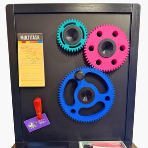 May include: A black wooden chalkboard with three colorful gear-shaped magnets in teal, pink, and blue. A yellow notepad with the word "MULTITASK" and a to-do list is attached. A red clip and a small tray with red pens are also visible.