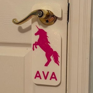 May include: A white door hanger with a pink unicorn silhouette and the name "AVA" printed in pink letters.
