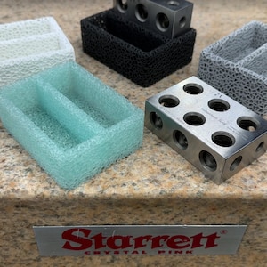 May include: A set of four metal blocks with different sized holes, used for precision measurement. The blocks are sitting on a granite surface with a Starrett Crystal Pink label.