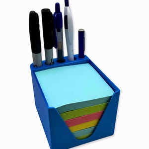 May include: A blue desk organizer with a stack of colorful sticky notes and several pens and markers. The organizer has a rectangular shape with a cutout on one side for easy access to the notes.