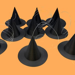 May include: A collection of small, black witch hats with a conical shape and wide brims. The hats are arranged on an orange background, with thin, white, looping strings intertwined among them. A festive Halloween theme.