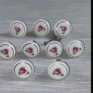 May include: A set of ten white ceramic cabinet knobs with a gold trim and a pink rose design. The knobs are round and include the screws for installation. Ideal for adding a touch of vintage charm to furniture.