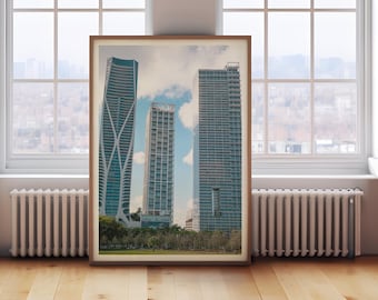 Può includere: Una stampa incorniciata con un paesaggio urbano con tre grattacieli moderni contro un cielo nuvoloso. Gli edifici sono di varie altezze e design, con un parco verde visibile alla base. La cornice è di colore marrone caldo e la stampa è esposta su un pavimento in legno.