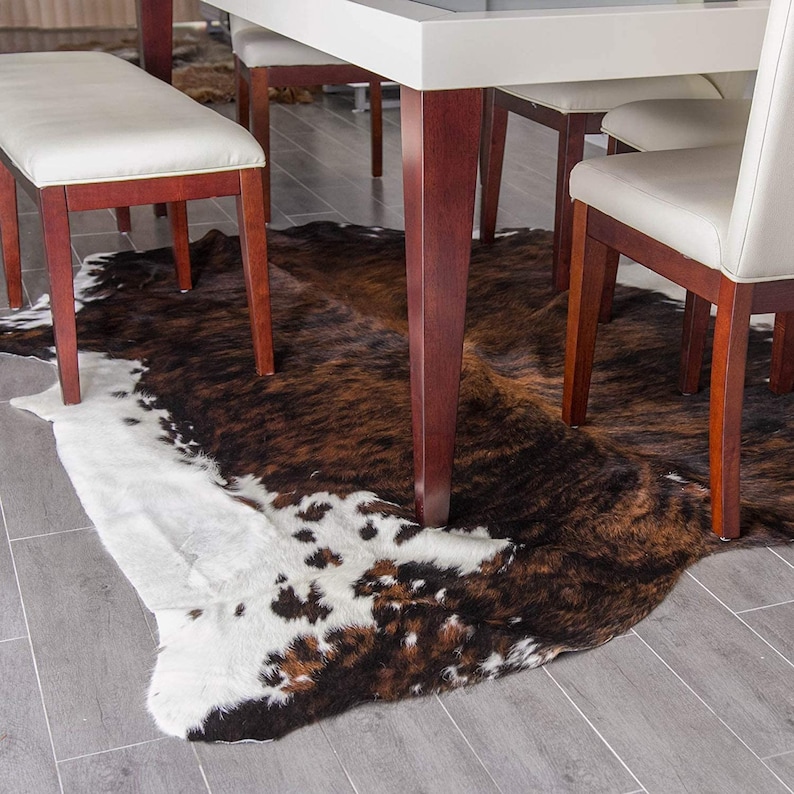 May include: A cowhide rug with a natural pattern of brown, black, and white. The rug is placed under a white dining table and chairs with dark wood legs. The rug's irregular shape adds a rustic touch to the dining area.