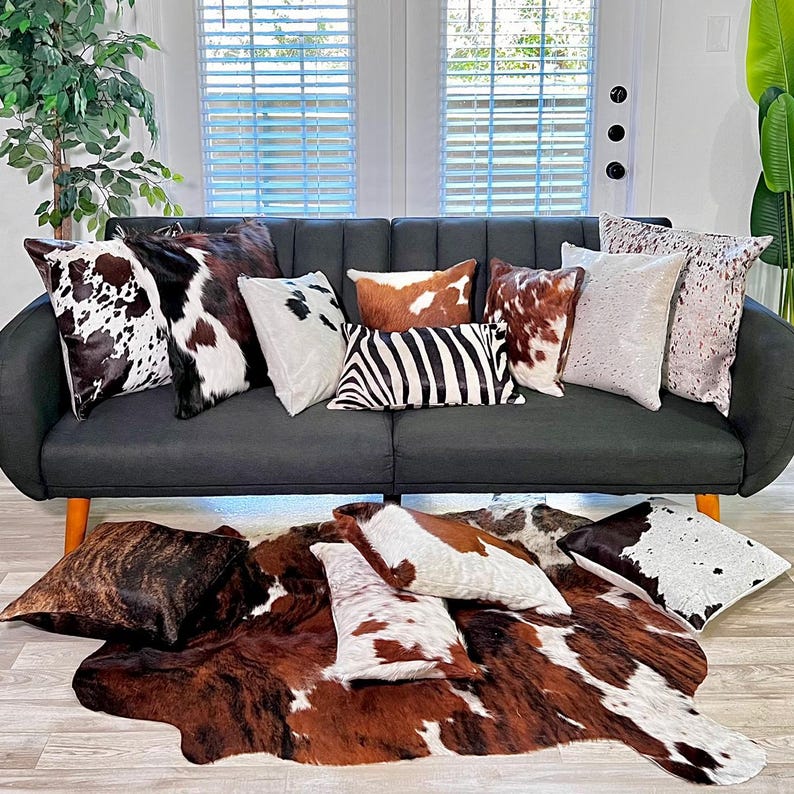 May include: A grey sofa with a variety of cowhide and zebra print cushions. The sofa is sitting on a light wood floor with a large cowhide rug in front of it.