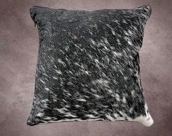 May include: Square decorative pillow with a cowhide pattern. The pillow features a black and white spotted design, with a zipper closure along the top edge. The pillow is designed for home decor.