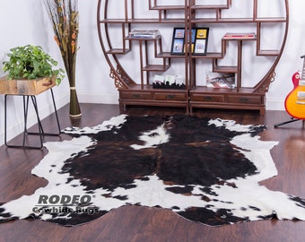 May include: A black and white cowhide rug with brown patches. The rug is laid out on a wooden floor in a room with a wooden shelf unit and a guitar stand with a guitar on it.