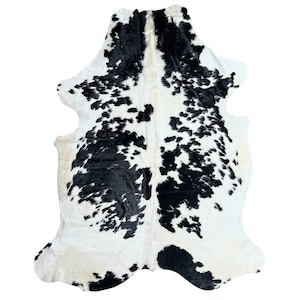 Puede incluir: Una alfombra de piel de vaca con un patrón natural de manchas negras sobre un fondo blanco. La alfombra tiene una forma irregular, con bordes redondeados y un aspecto suave y texturizado. Esta alfombra decorativa es adecuada para la decoración del hogar.