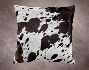 May include: Square decorative pillow with a cowhide pattern. The pillow features a white background with irregular black spots and patches. The pillow has a soft, textured appearance and is likely used for home decor.