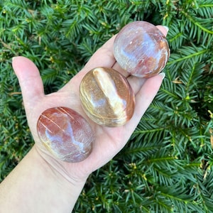 May include: Three smooth, polished, red and white striped, oval-shaped stones. The stones are held in a hand.