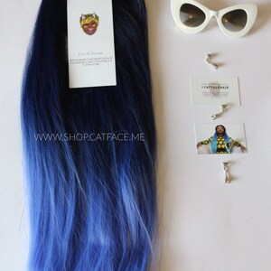 May include: A long, straight, ombre hair extension with dark blue at the top and light blue at the bottom. The hair extension is on a white background with a business card that says "#CATFACHAIR" and a pair of white cat-eye sunglasses.
