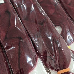 Puede incluir: Múltiples paquetes de pelo trenzado burdeos, cada uno con una banda elástica en el extremo. Los paquetes están etiquetados como "Catface Hair" y "Braiding Hair".