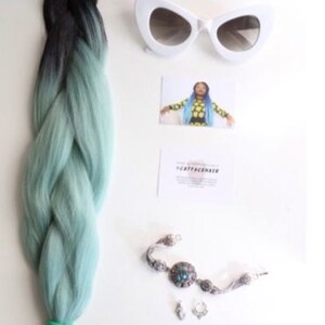 May include: A braid of black and light blue hair extensions. The hair is braided and has a ombre effect. The hair is on a white background with a pair of white cat eye sunglasses, a small card with the text "#CATFACEHAIR", a silver bracelet with a turquoise stone, and two silver earrings.