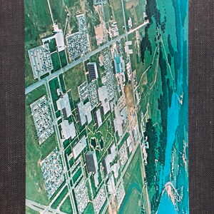 May include: An aerial view of a large industrial complex with multiple buildings, parking lots, and green spaces. The complex is surrounded by trees and a body of water.