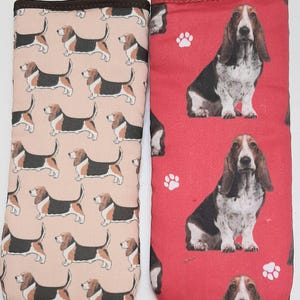 Puede incluir: Dos manoplas para horno con un patrón de perro de caza. Una manopla es rosa con perros de caza negros y la otra es roja con huellas de patas blancas y perros de caza negros y blancos.