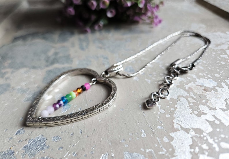 May include: A silver heart-shaped pendant necklace with a rainbow of small beads hanging inside the heart. The necklace has a silver chain and a lobster clasp.