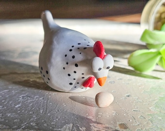 Peut inclure: Une figurine de poulet en céramique blanche avec des pois noirs, une crête et des barbillons rouges, et un bec orange, regardant un petit œuf clair. La figurine est sur une surface texturée, suggérant un thème de ferme ou de cuisine.