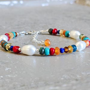 May include: A colorful beaded bracelet featuring a variety of gemstones and large white pearls. The bracelet includes red, blue, orange, and green beads, connected by a silver chain. A stylish accessory.
