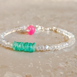 May include: A delicate bracelet with clear, faceted beads. The bracelet is accented with a vibrant pink bead and a section of bright green beads. The clasp is silver-toned. The bracelet is displayed on a neutral surface.
