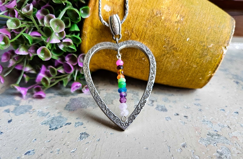 May include: A silver heart-shaped pendant necklace with a rainbow of gemstone beads hanging inside. The pendant has an ornate design around the edge.