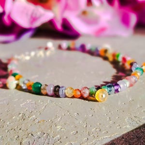 May include: A colourful bracelet made of small beads in shades of green, orange, purple, and yellow. The bracelet is strung on a thin, silver-coloured chain. The background features blurred pink and purple flowers.