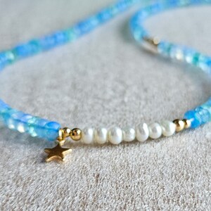 Peut inclure: Un collier d&eacute;licat en perles bleues et blanches avec un pendentif &eacute;toile en or. Le collier est compos&eacute; de petites perles rondes et comporte une rang&eacute;e de perles blanches.