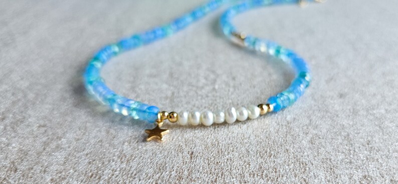 Peut inclure: Un collier d&eacute;licat en perles bleues et blanches avec un pendentif &eacute;toile en or. Le collier est compos&eacute; de petites perles rondes et comporte une rang&eacute;e de perles blanches.