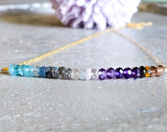 May include: A delicate gold chain necklace featuring a row of colorful gemstone beads. The beads transition from light blue to purple and brown. The necklace is displayed on a light gray surface.