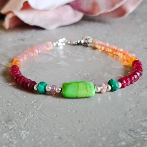 May include: A beaded bracelet featuring a central rectangular lime green stone, flanked by small round green and pink beads, faceted deep red beads, and translucent orange beads, all accented with small silver spacers and a silver clasp closure.