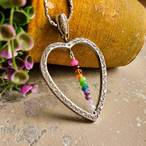 May include: A silver heart-shaped pendant necklace with a rainbow of gemstone beads hanging inside. The pendant has an intricate design and the beads are colourful and vibrant.