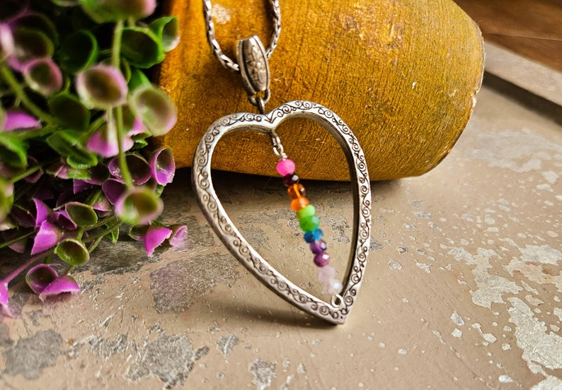 May include: A silver heart-shaped pendant necklace with a rainbow of gemstone beads hanging inside. The pendant has an intricate design and the beads are colourful and vibrant.