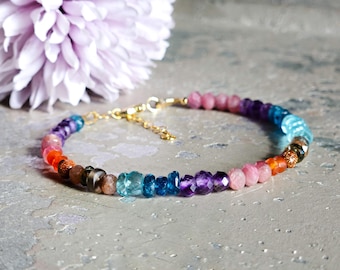 May include: A colorful beaded bracelet with a gold clasp. The bracelet features a variety of gemstones in shades of pink, orange, blue, and purple. A light purple flower is in the background.