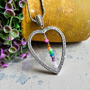 May include: A silver heart-shaped pendant necklace with a rainbow of gemstone beads hanging inside. The necklace is on a silver chain.