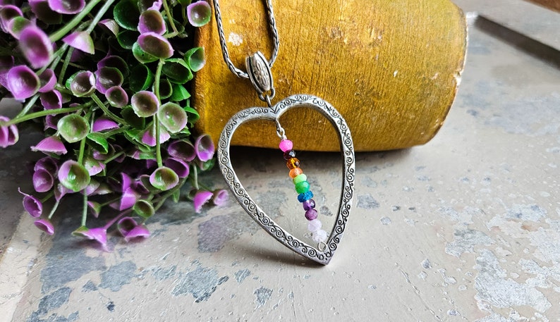 May include: A silver heart-shaped pendant necklace with a rainbow of gemstone beads hanging inside. The necklace is on a silver chain.
