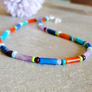 May include: A colorful beaded necklace with a variety of cylindrical and round beads. The beads are in shades of blue, orange, purple, green, and white. The necklace has a silver clasp and is displayed on a light-colored surface.